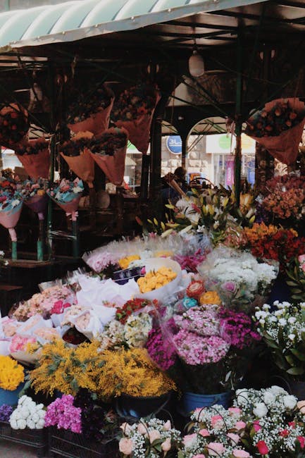 A vibrant floral display featuring a variety of fresh flowers arranged in bouquets and loose stems, including predominantly white, yellow, pink, and purple blooms such as roses, chrysanthemums, and ba