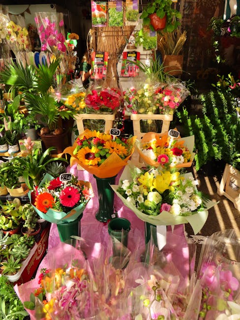 A vibrant display of fresh floral arrangements and bouquets arranged on a pink cloth-draped table inside a florist shop. The bouquets feature a variety of flowers including bright red, yellow, white, 