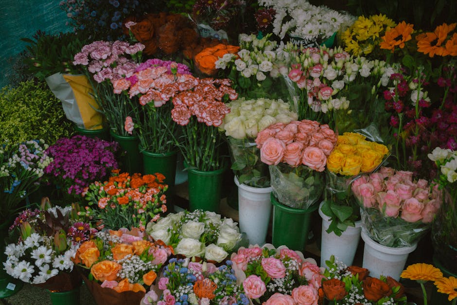 A vibrant display of fresh flowers arranged in various bouquets and individual stems, featuring roses in soft pink, white, and yellow, alongside bright orange and purple blooms, all wrapped in clear o