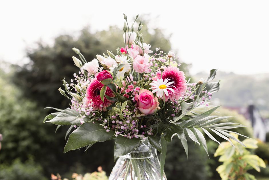 A fresh flower bouquet featuring pink roses, white daisies with yellow centers, pink gerbera daisies, and small pink filler flowers, arranged with lush green foliage and large palm leaves in a clear g