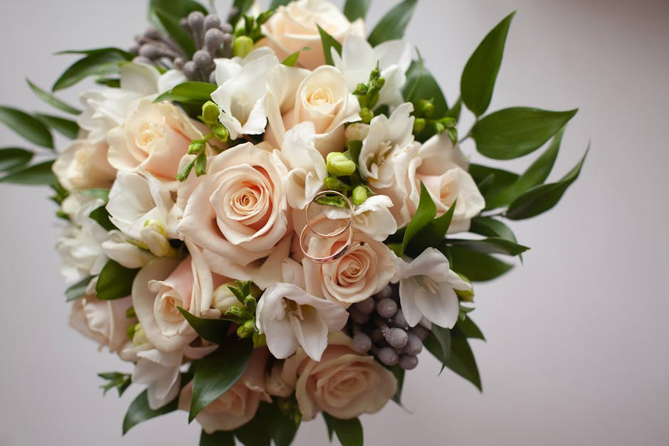 A close-up view of a floral bouquet featuring pale pink roses with tightly furled petals and creamy white freesias with delicate, trumpet-shaped blossoms. The arrangement is accented with small green 