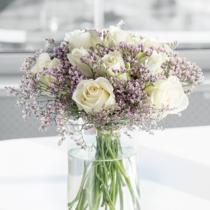 A beautifully composed arrangement of sympathy flowers fills the frame: approximately a dozen creamy white roses and a soft cloud of lavender-pink filler blossoms, carefully gathered in a tall, clear glass vase. Each rose is in full bloom, its petals gently unfurling in thick, velvety layers that cradle a classic spiral heart, their colour shifting from pure white at the outer edges to a subtle ivory glow near the center. Threaded among the roses are plentiful sprigs of delicate statice or limonium, their tiny flowers forming feathery clusters in tones of pale lilac and blush, lending the design a light, airy texture. The stems, neatly trimmed and vibrant green, are visible through the cylindrical vase, extending down into the transparent water that holds them upright, suggesting the freshness and care that went into this arrangement. The vase rests on a pristine white surface, perhaps a side table in a quiet South Wimbledon home, its gloss catching echoes of natural light. Behind the bouquet, the background is gently blurred in shades of white and soft grey, evoking the calming atmosphere of a peaceful room near Wimbledon Common or a tranquil corner of a local chapel. There is no clutter to distract from the flowers; instead, the composition speaks softly of remembrance, dignity, and tender support. Created in the style of Florist South Wimbledon's thoughtful tributes, this bouquet feels ideal for expressing condolences, whether delivered to a bereaved family's door or placed beside a framed photograph during an intimate memorial gathering.