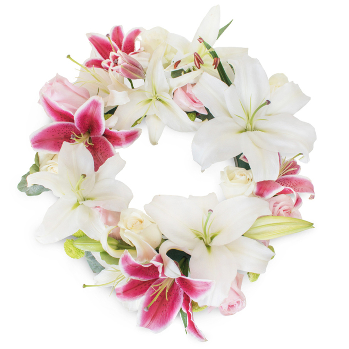 Centered in the calm elegance of South Wimbledon, this sophisticated floral wreath presents an exquisite blend of traditional beauty and modern sensitivity. Its full, unbroken circle signifies continuity and eternal memory, rendered in a dramatic mix of snowy white lilies and vivid fuchsia pink lilies. The star-like forms of the white lilies dominate the composition, their unfurled petals and prominent green pistils recalling peace and gentle reflection. Scattered throughout, bursts of fuchsia lilies announce themselves with confident color-a deep magenta touched with faint speckles-introducing vibrant contrast and channeling cherished memories. Complimenting these lilies, you'll notice clusters of blushing roses in both soft pink and cream shades. Their petals are luxuriously dense, some tightly rolled, others blossoming open, adding layers of texture and embracing the arrangement in romantic warmth. Wisps of fresh greenery, possibly echoing the foliage found strolling through nearby Morden Hall Park, offer quiet balance and natural depth, preventing the floral ring from feeling heavy-massed. Set against a crisp white background, every detail is beautifully accentuated, allowing the colors and shapes to speak their message of sympathy and hope. This memorial wreath, expertly created by Florist South Wimbledon, provides a graceful tribute for funerals or celebrations of life, ensuring every gesture of condolence is wrapped in beauty and sincerity.