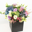 Presented against a clean white backdrop, this radiant and sophisticated bouquet from South Wimbledon is a visual feast of colour, texture, and artistry. A matte black square bag grounds the lush composition, elevating every blossom within. Dominating the centre are sumptuous, billowy hydrangeas in subtle shades of deep blue, indigo, and silvery mauve, their petals plush and velvety. Nestled alongside them, tightly coiled roses in soothing lavender and gentle blush tones offer a romantic flourish, their delicate folds catching the daylight familiar to quiet residential streets nearby. Creamy lisianthus, with trumpet-like blossoms in pure white and buttery yellow, rise elegantly, adding airiness and a feeling of freshness. Lustrous lime green dianthus balls introduce playful texture, reminiscent of herbaceous borders in local parks. Soft sprays of feathery pink astilbe weave between the larger flowers, introducing graceful movement and an almost whimsical charm. Interspersed greenery and fresh green buds peek through the abundance, giving the piece natural vibrancy and balance. The whole arrangement feels modern, inviting, and luxurious-perfect for marking heartfelt celebrations, expressing gratitude, or beautifying a South Wimbledon home or office. This floral design, expertly crafted and available for delivery throughout South Wimbledon, captures the local spirit and attention to detail that make the area's florists renowned for their bespoke creations.